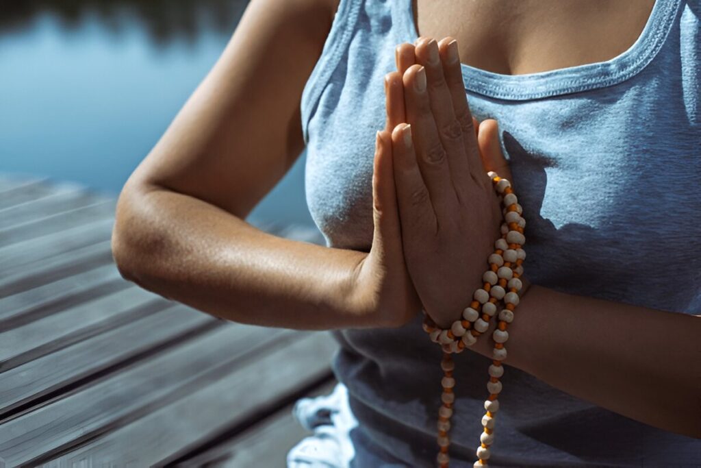 The young woman sitting on a wooden pier with her hands folded with a rosary in a prayer pose. Meditation, yoga in nature. Close-up.